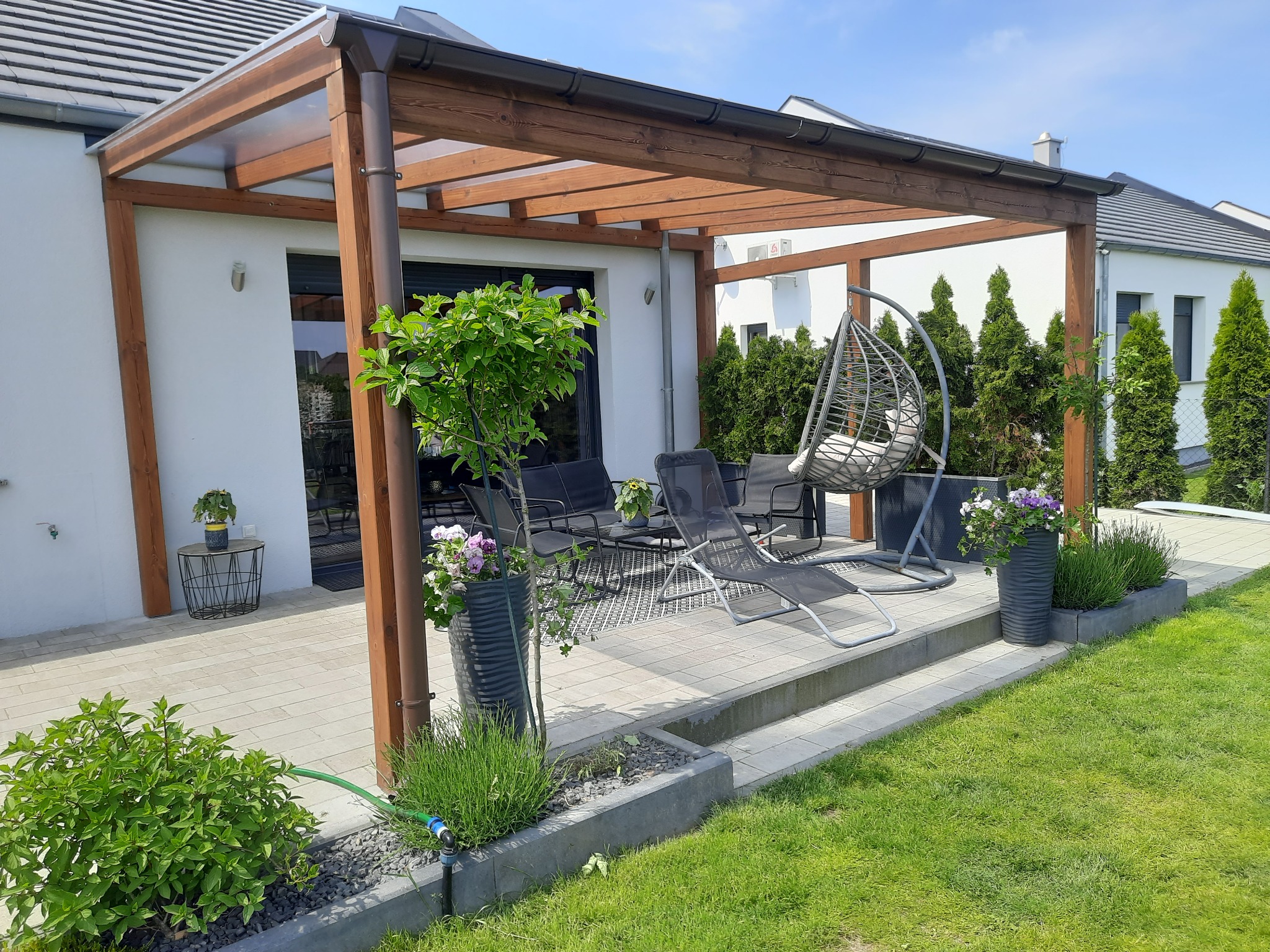 Zadaszony taras z drewnianą konstrukcją, wyposażony w meble wypoczynkowe, wiszący fotel i rośliny w donicach, otoczony zadbanym trawnikiem.