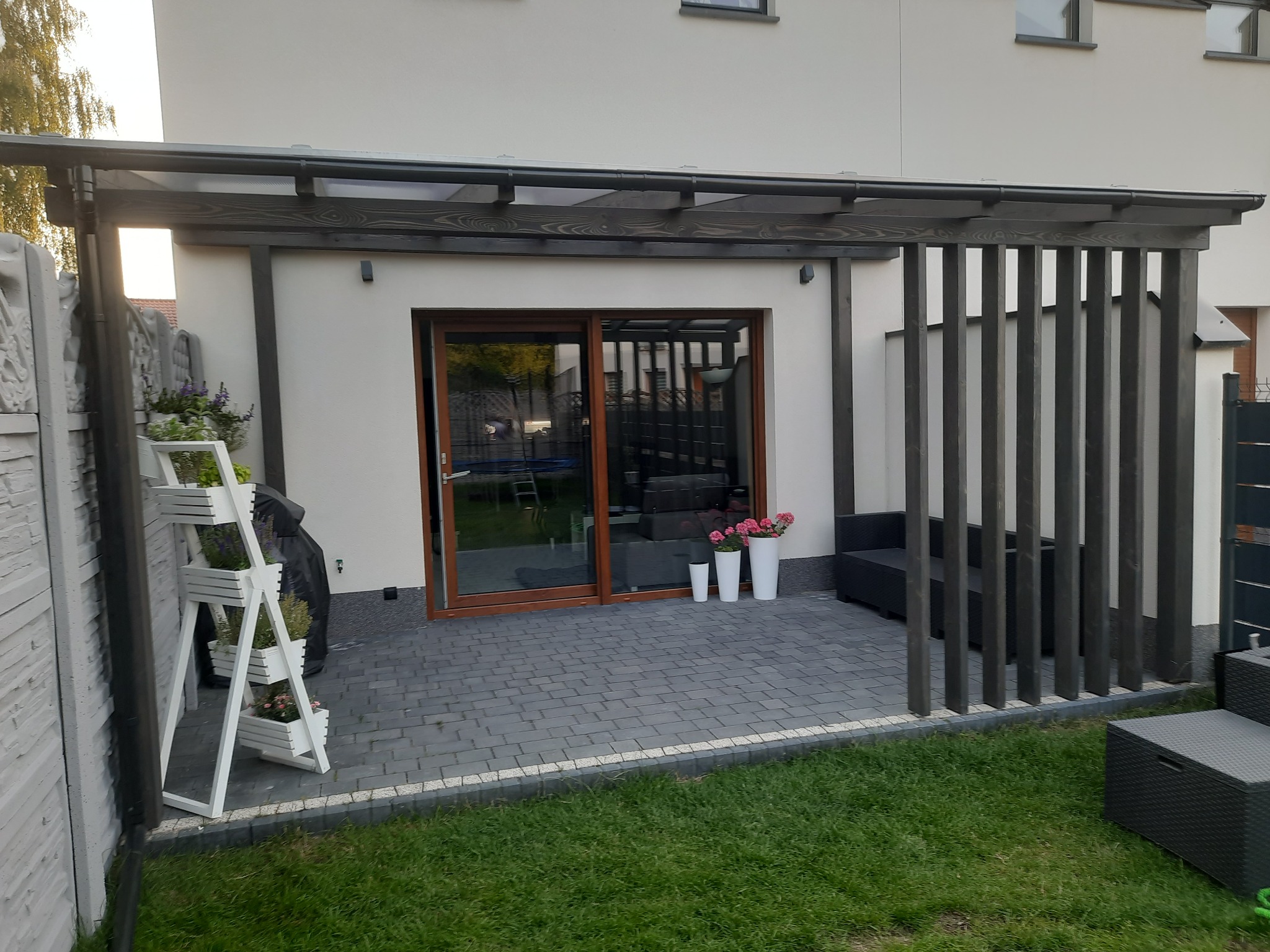 Nowoczesna pergola drewniana z zadaszeniem i pionowymi panelami w Lesznie, przylegająca do domu z wyjściem na taras wyłożony kostką, uzupełniona o dekoracje roślinne i meble ogrodowe.