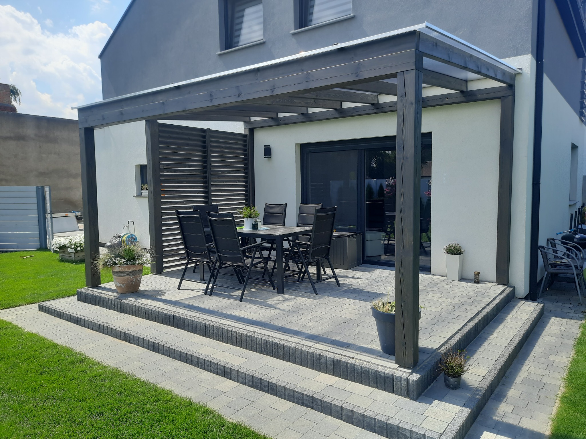 Nowoczesna pergola z ciemnego drewna z przezroczystym zadaszeniem, z kompletem mebli ogrodowych na tarasie z jasnoszarej kostki, trzy stopnie schodkowe, trawnik i kwiaty w donicach.