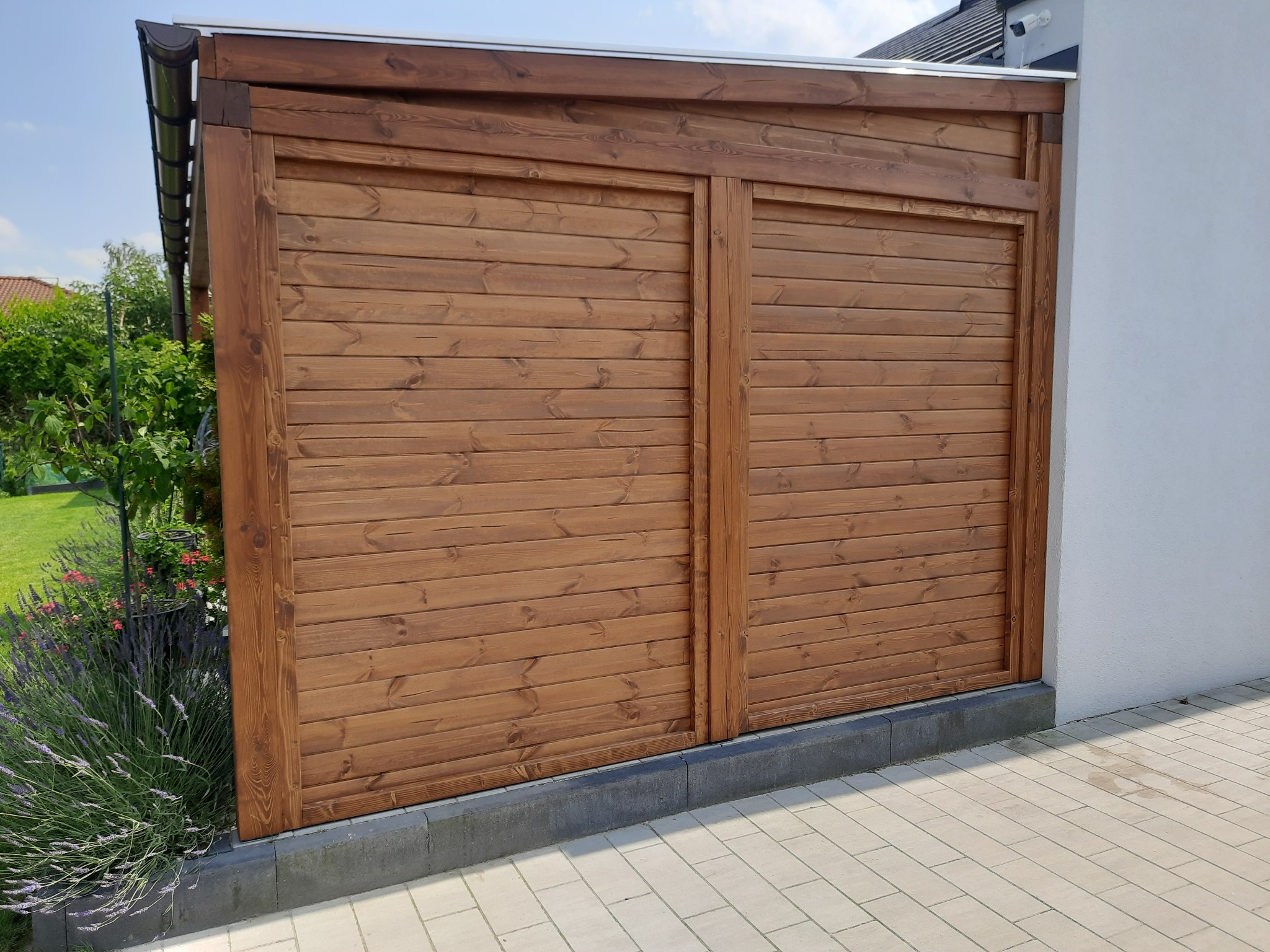 Drewniana konstrukcja z poziomych desek, przylegająca do białej ściany budynku, z widocznym fragmentem dachu i zielenią w tle.