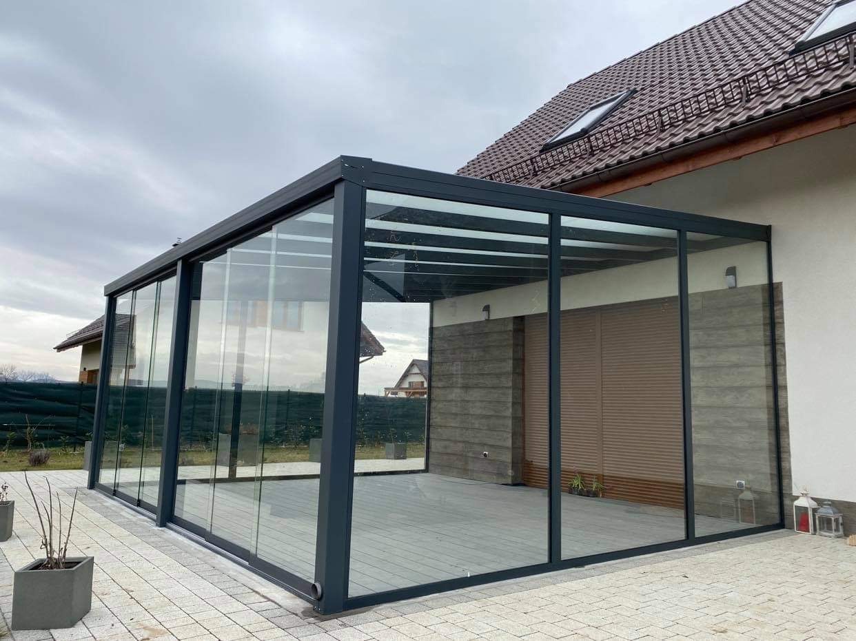Nowoczesna, przeszklona konstrukcja z ciemną ramą, prawdopodobnie metalowa, przylegająca do budynku z brązową dachówką; widoczne przesuwne szklane panele i szara nawierzchnia wewnątrz.