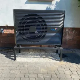 Czarna pompa ciepła marki FOX Air na metalowym stelażu, ustawiona na białym żwirku, przed ścianą z cegły i białego tynku.