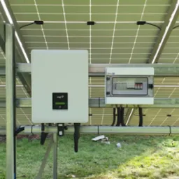 Widok od dołu na panele słoneczne, falownik marki Fox ESS i skrzynkę zabezpieczeń na tle zielonego trawnika i tui.