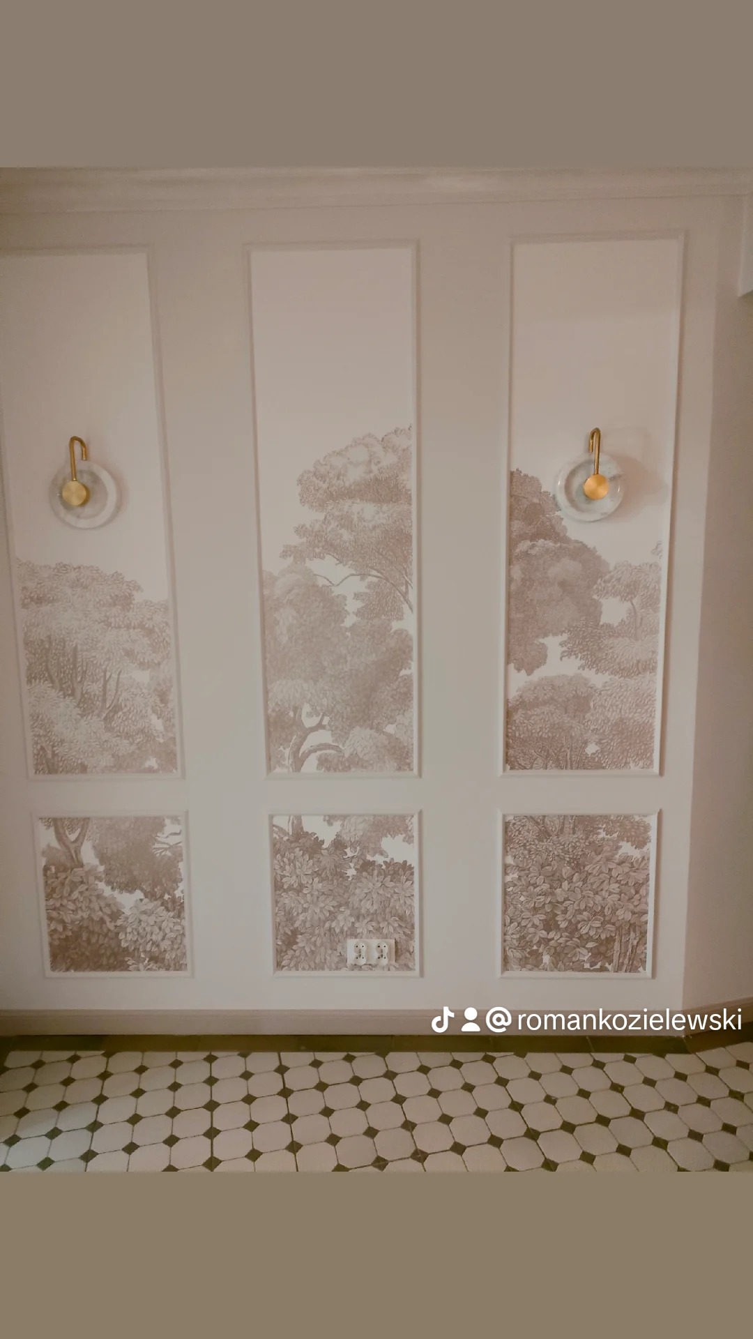 Tapeta z motywem drzew w odcieniach beżu, podzielona na panele na ścianie, uzupełniona o kinkiety z marmurowymi podstawami i złotymi uchwytami, nad podłogą w czarno-białą mozaikę.