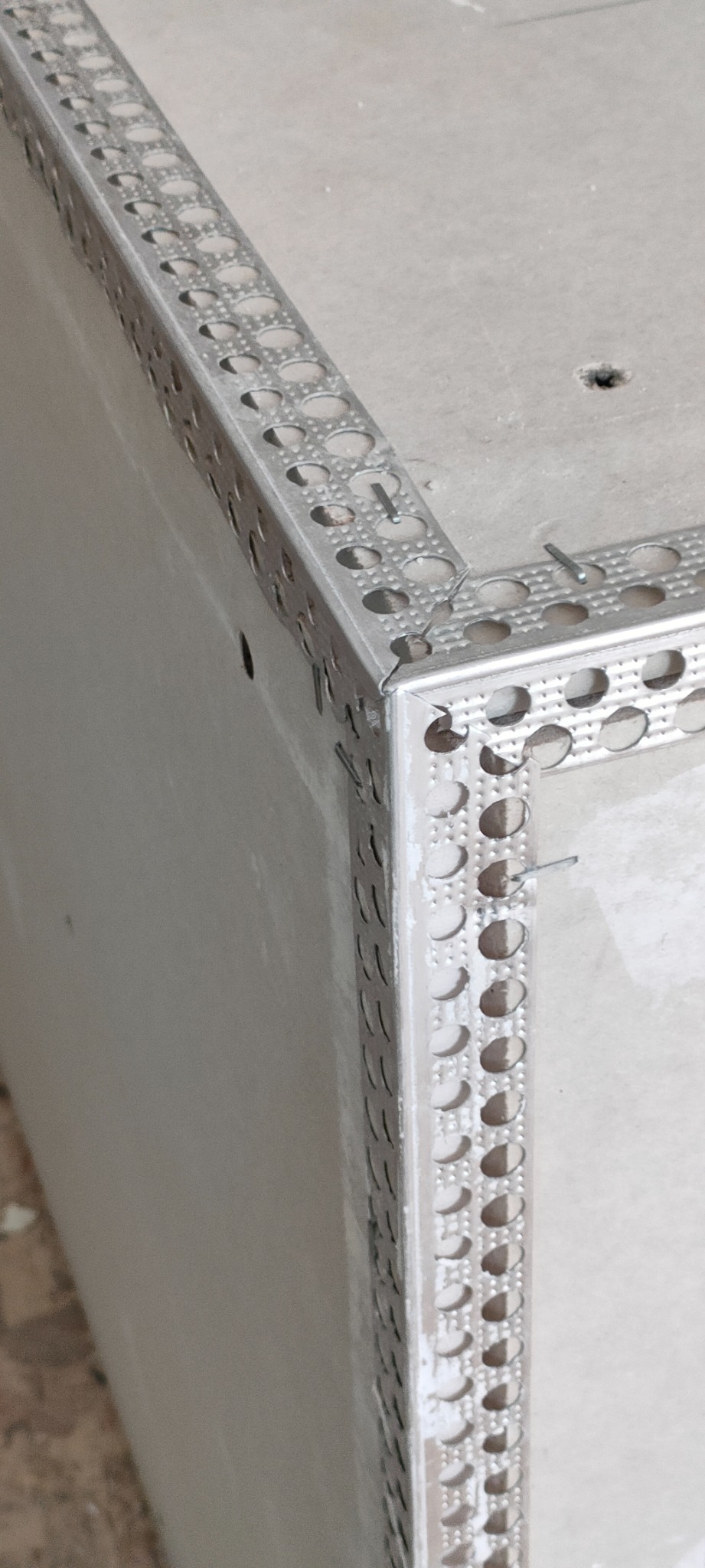 Metalowy narożnik perforowany przymocowany do ściany gwoździami, przygotowany pod wykończenie.