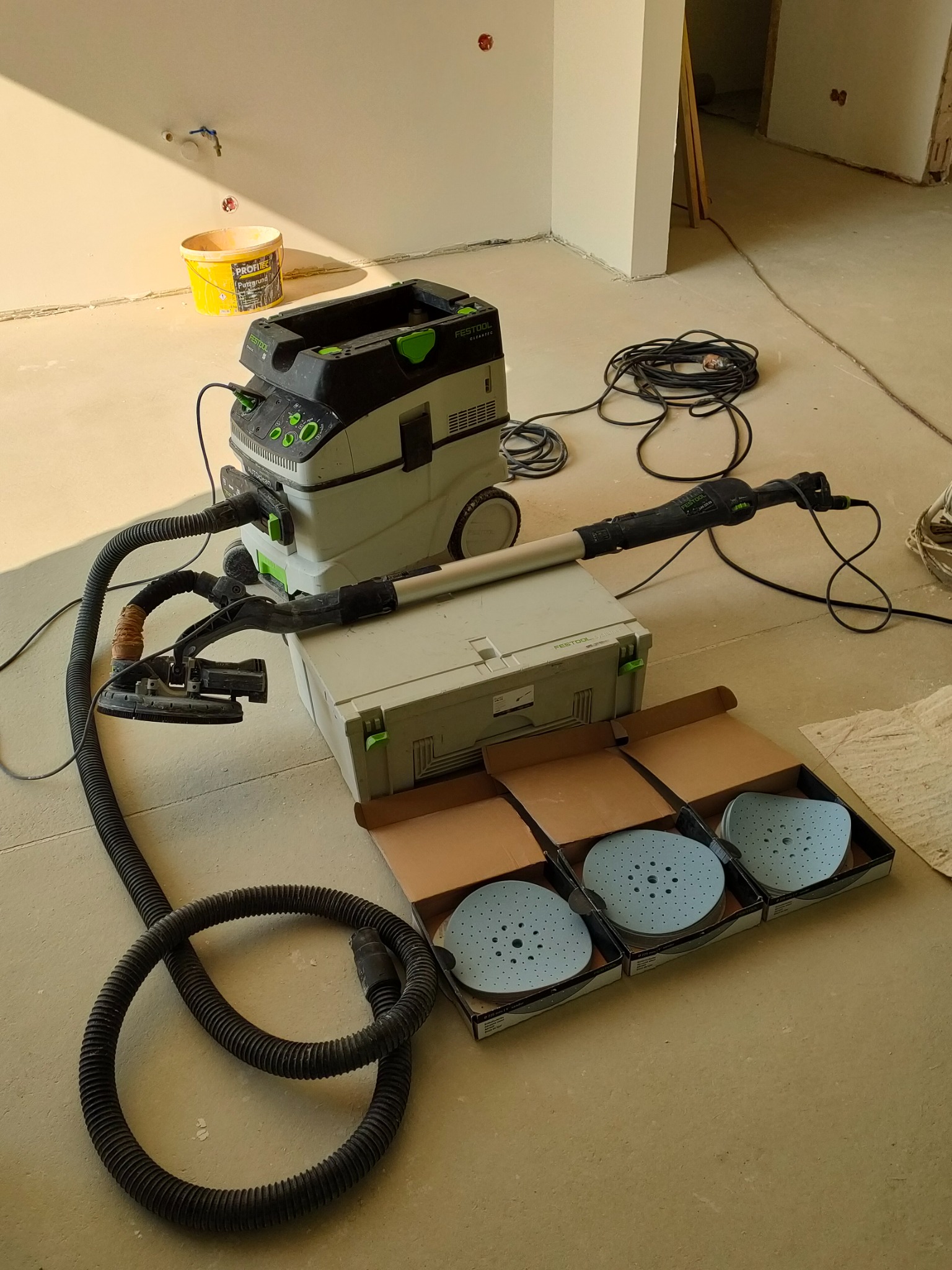 Szlifierka do gładzi na wysięgniku Festool Planex LHS 225 z odkurzaczem i zestawem tarcz szlifierskich w otwartych opakowaniach, na tle surowego wnętrza w trakcie prac wykończeniowych.