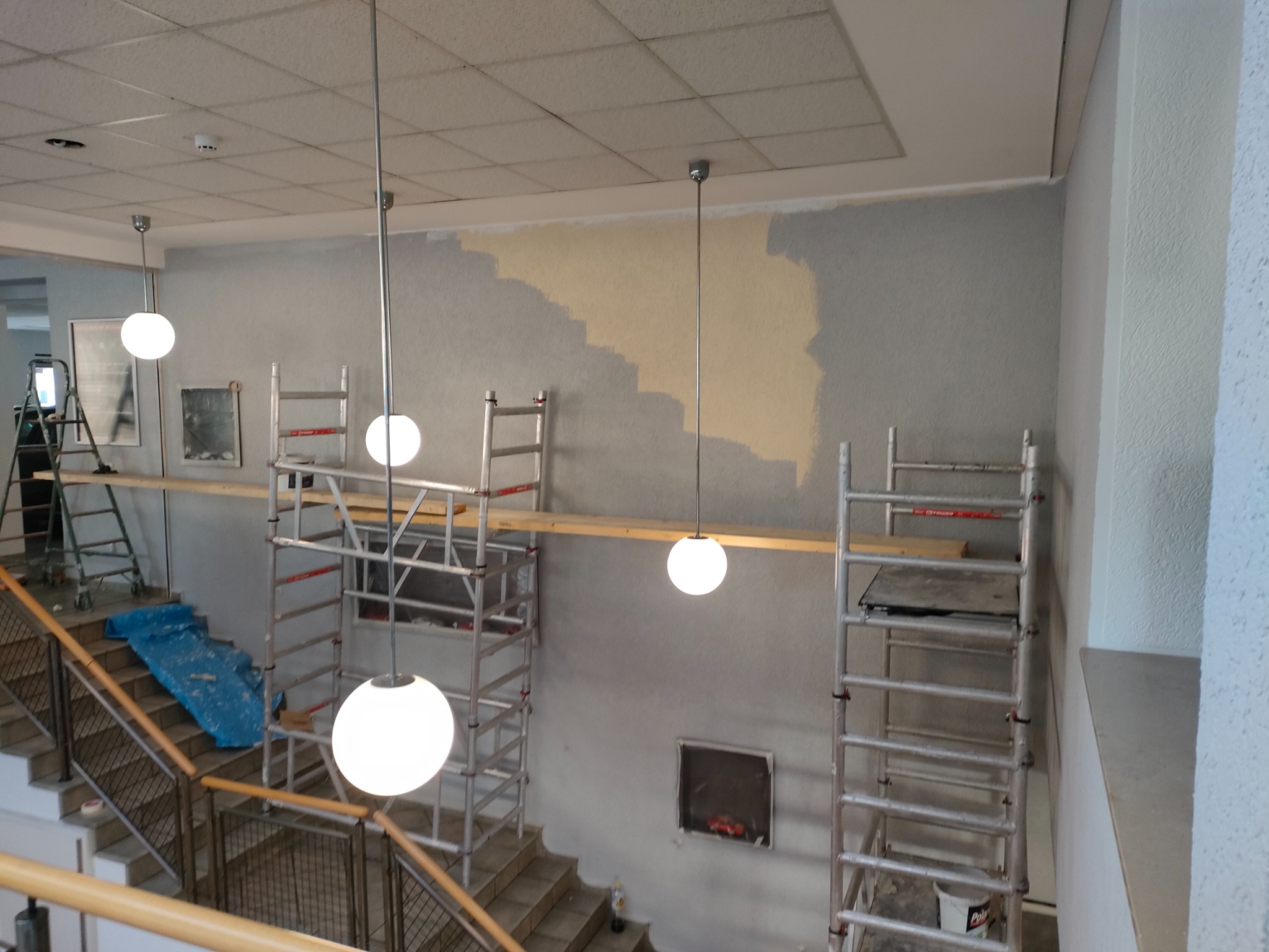 Wnętrze klatki schodowej w trakcie malowania, widoczne rusztowania, fragment pomalowanej ściany na beżowo oraz niebieska folia zabezpieczająca schody.