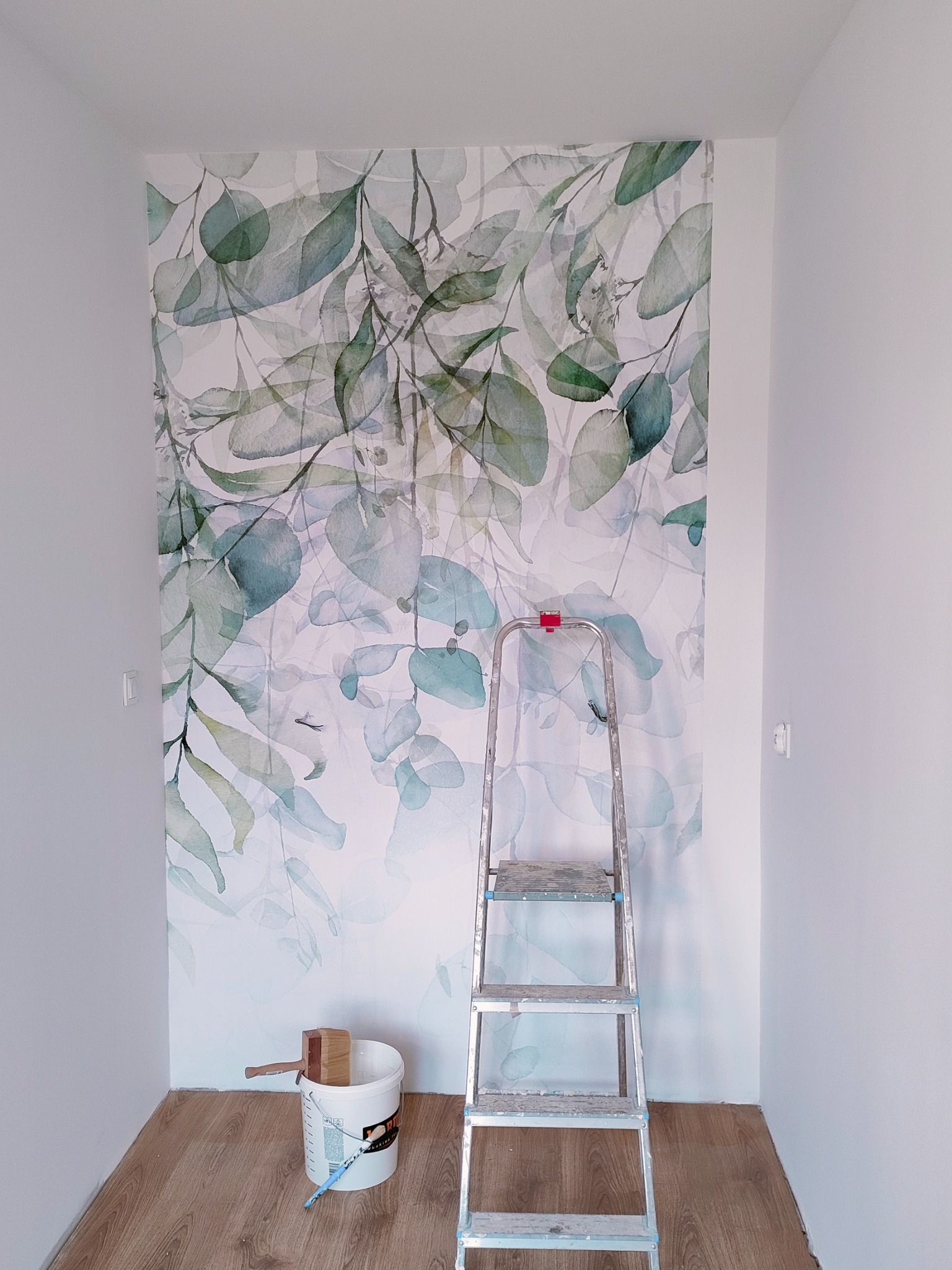 Fragment pokoju po położeniu tapety z motywem akwarelowych liści eukaliptusa, widoczna drabina i wiadro z narzędziami.