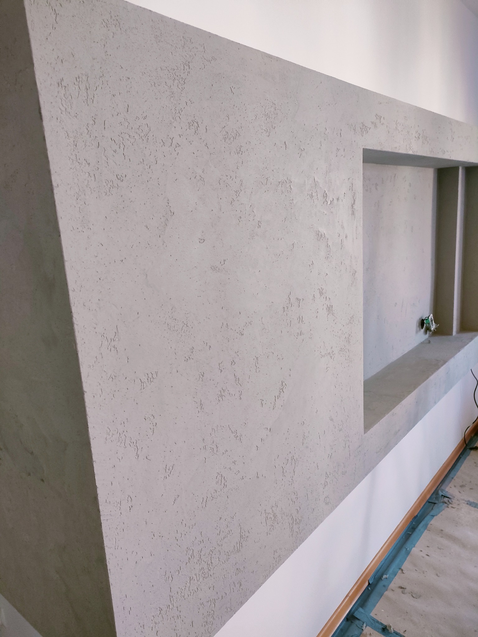 Fragment ściany z szarą dekoracyjną strukturą, wnęka z półkami i widoczne wykończenie podłogi w trakcie prac.
