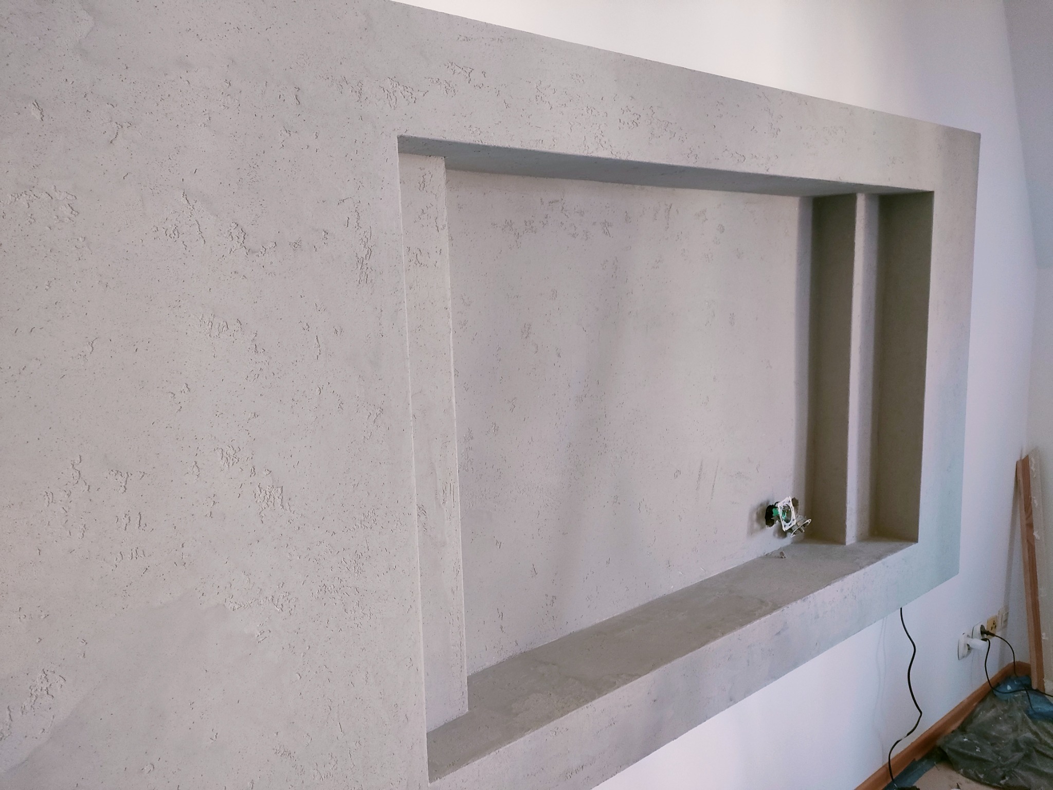 Ściana z szarą, teksturowaną strukturą, wnęka w ścianie z widocznym gniazdkiem elektrycznym, surowy stan wykończenia.