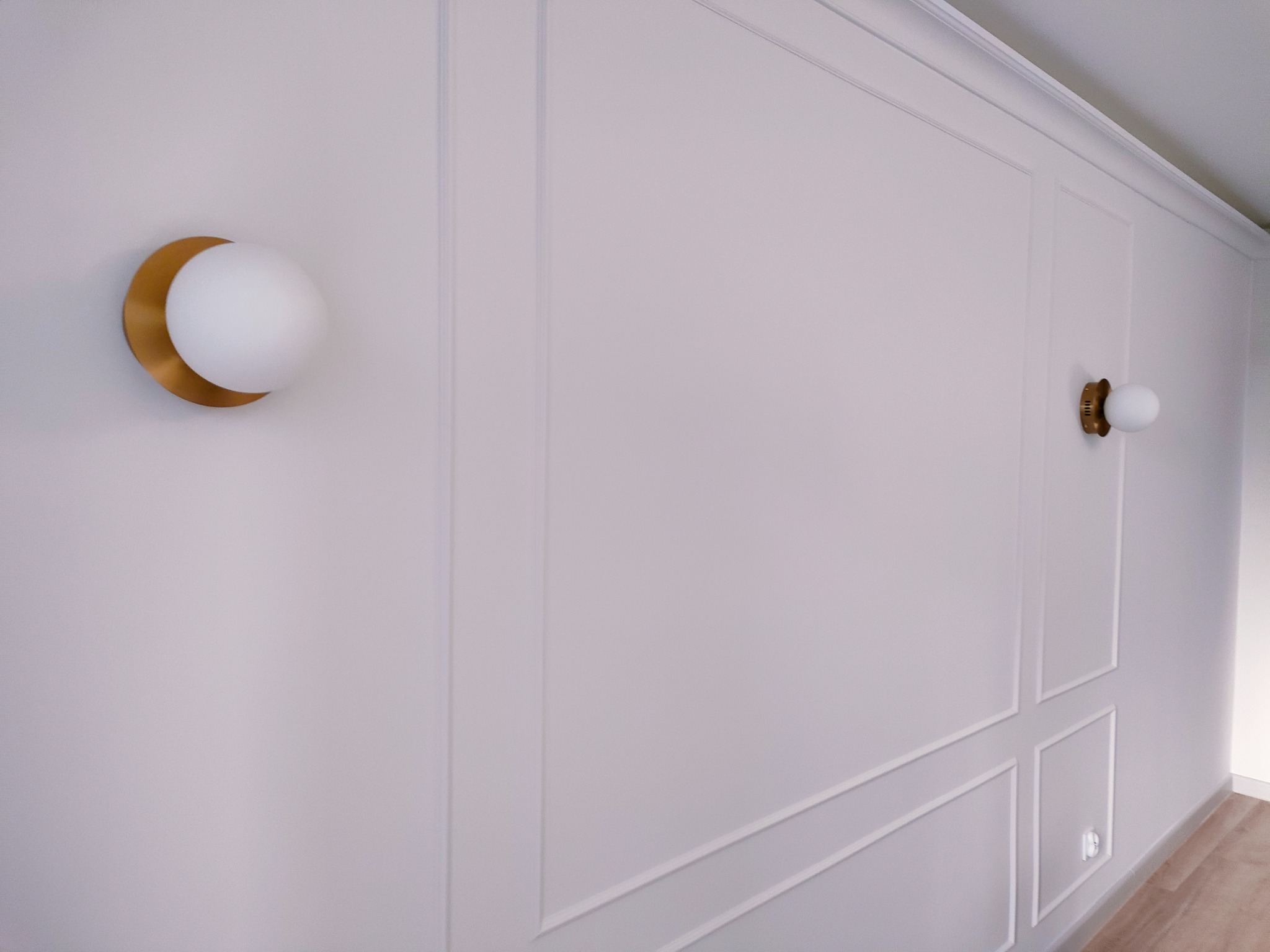 Ściana z białymi panelami dekoracyjnymi i dwoma nowoczesnymi lampami ściennymi z kulistymi kloszami i złotymi elementami mocującymi.