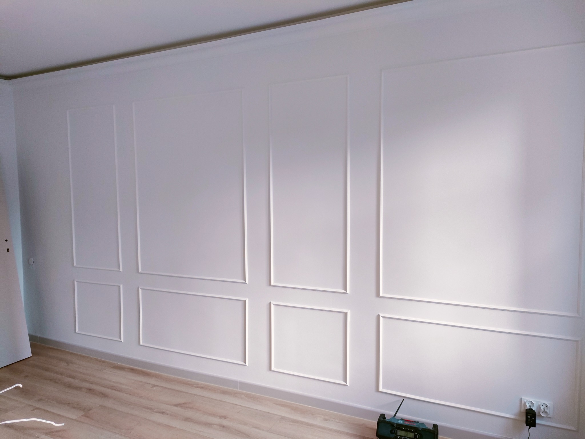 Biała ściana z dekoracyjnymi listwami w kształcie prostokątów, jasne panele podłogowe, widoczny fragment drzwi i sprzęt audio na podłodze.