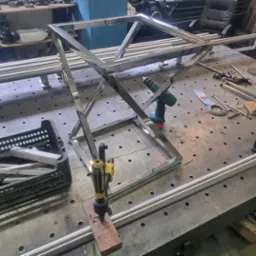 Warsztatowy stół z perforowanym blatem, na którym znajdują się elementy konstrukcji metalowej w trakcie montażu, w tle widoczna spawarka i inne narzędzia.