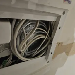 Otwarty panel dostępowy do przestrzeni z gęsto upakowanymi kablami elektrycznymi i telekomunikacyjnymi, widoczna perforowana metalowa płyta w tle.