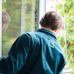 Mężczyzna w niebieskiej bluzie z logo na plecach reguluje mechanizm otwierania białego okna, z widocznym odbiciem w szybie i zielenią na zewnątrz.