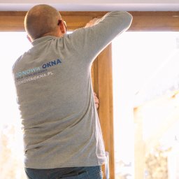 ODNOWA OKNA SPÓŁKA Z OGRANICZONĄ ODPOWIEDZIALNOŚCIĄ - Mężczyzna w szarej koszulce z logo firmy montuje okno z drewnopodobną ramą, widok od tyłu, jasne światło słoneczne wpadające przez okno.