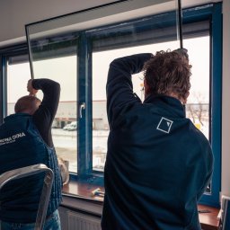 ODNOWA OKNA SPÓŁKA Z OGRANICZONĄ ODPOWIEDZIALNOŚCIĄ - Dwóch monterów w uniformach instaluje szybę w ramie okiennej; widok zza pleców, w tle budynek magazynowy i drabina.