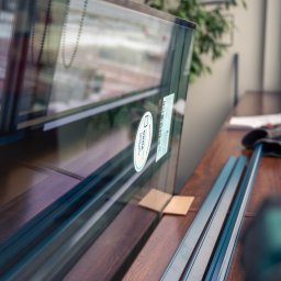 ODNOWA OKNA SPÓŁKA Z OGRANICZONĄ ODPOWIEDZIALNOŚCIĄ - Przygotowane do montażu okno z naklejkami producenta, leżące na drewnianym parapecie obok narzędzi i rękawic roboczych.