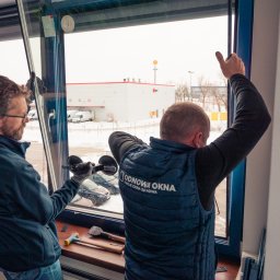 ODNOWA OKNA SPÓŁKA Z OGRANICZONĄ ODPOWIEDZIALNOŚCIĄ - Dwóch monterów instaluje okno, używając przyssawek do szkła; widoczne narzędzia i logo firmy na kamizelce jednego z nich; za oknem zimowy krajobraz z budynkami magazynowymi.