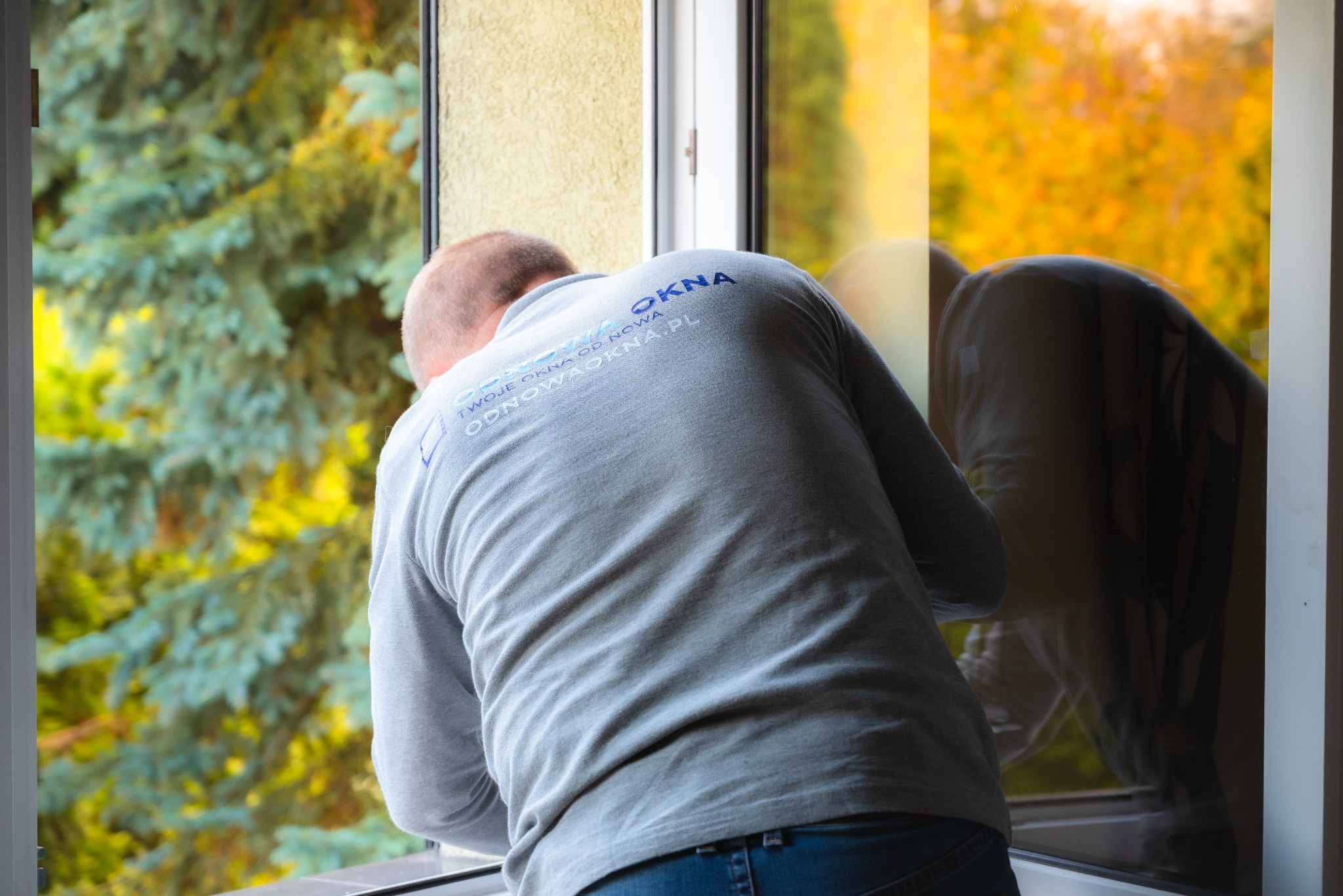Mężczyzna w szarej koszulce z logo firmy zajmującej się oknami, pochylony nad oknem, prawdopodobnie podczas montażu lub serwisu, z widokiem na zewnątrz.