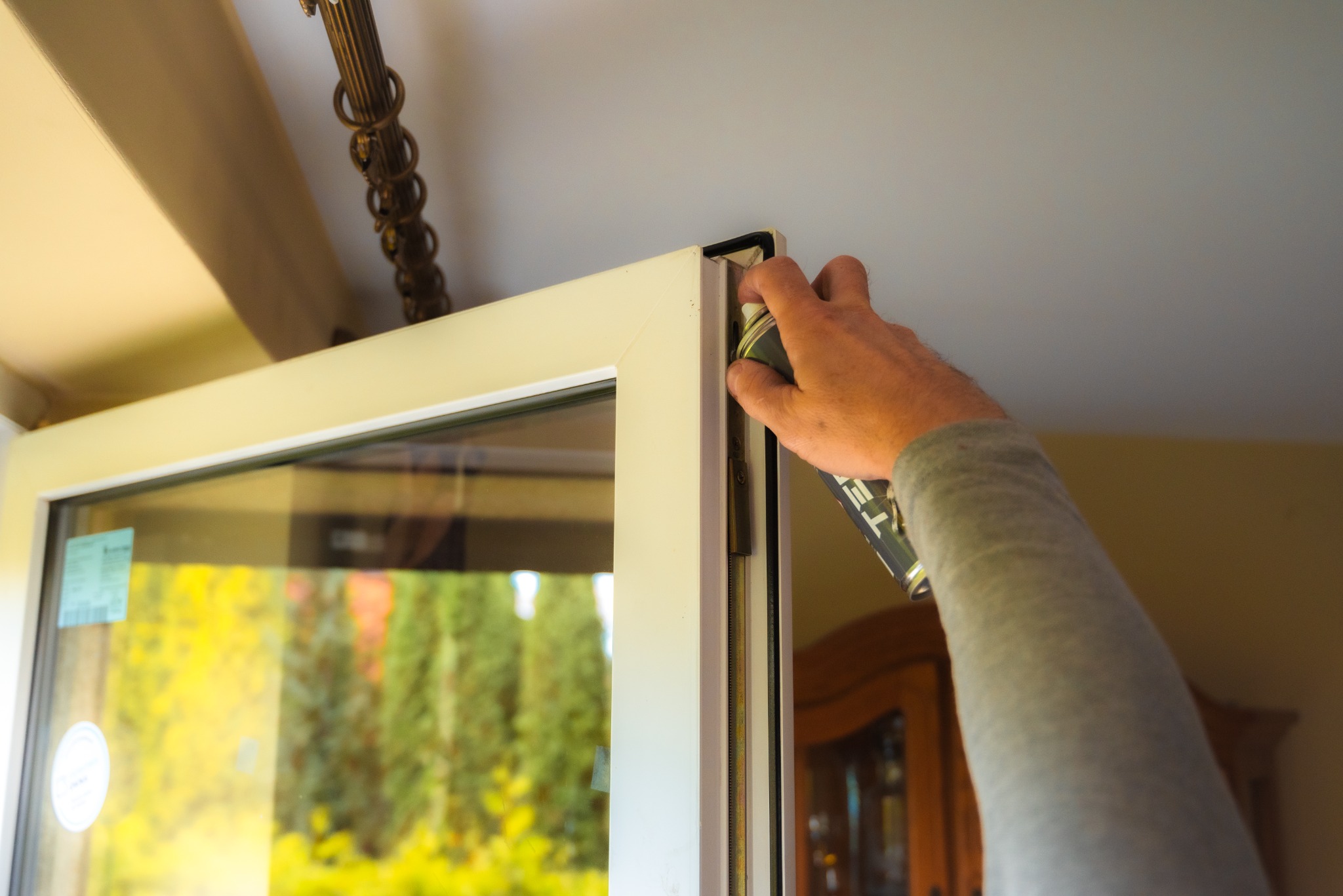 Malowanie uszczelki okna białą farbą w sprayu, widoczna rama okienna, szyba z widokiem na zieleń oraz fragment ozdobnego żyrandola.