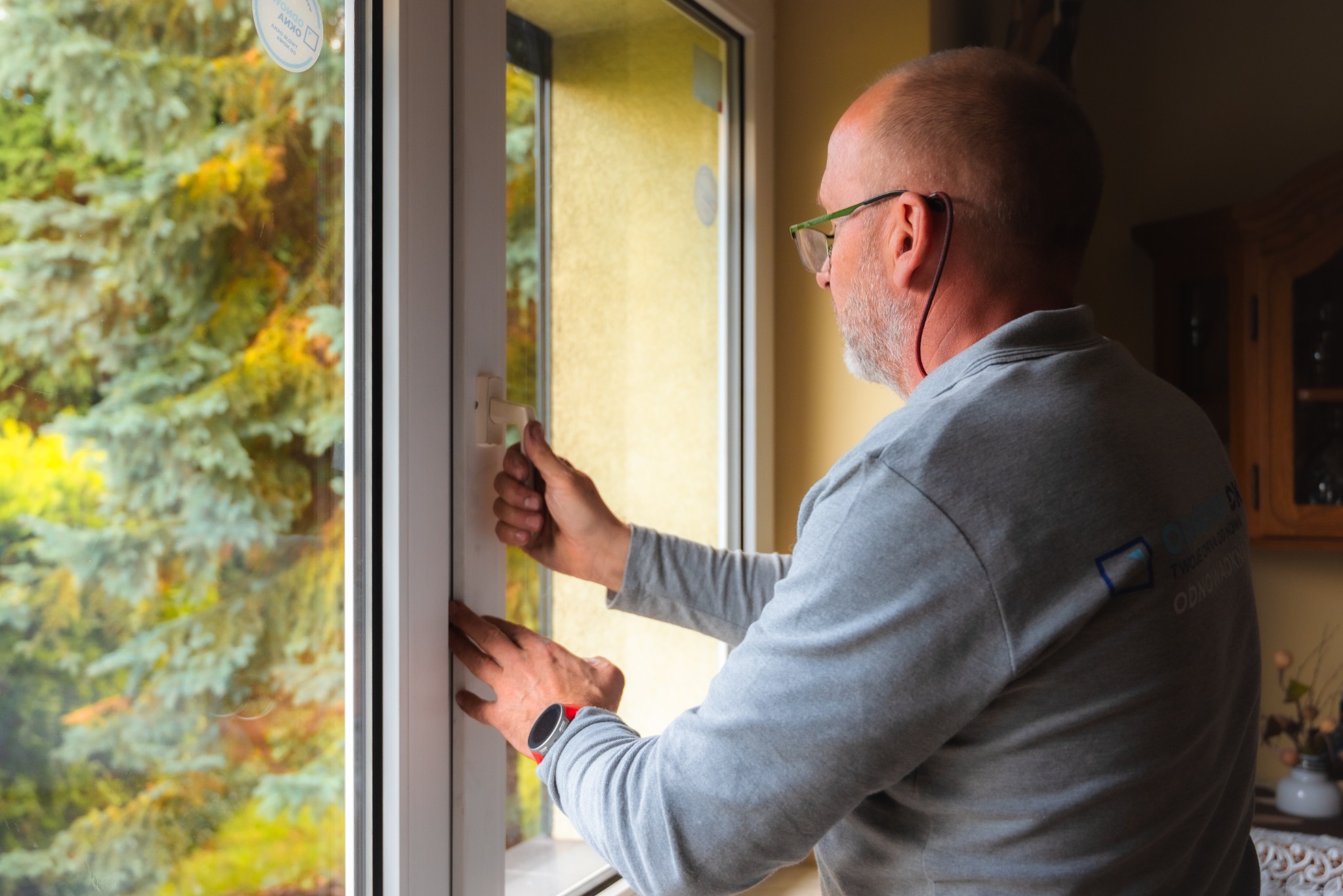 Mężczyzna w okularach reguluje białe okno PCV, widoczny profil okienny i krajobraz za oknem.