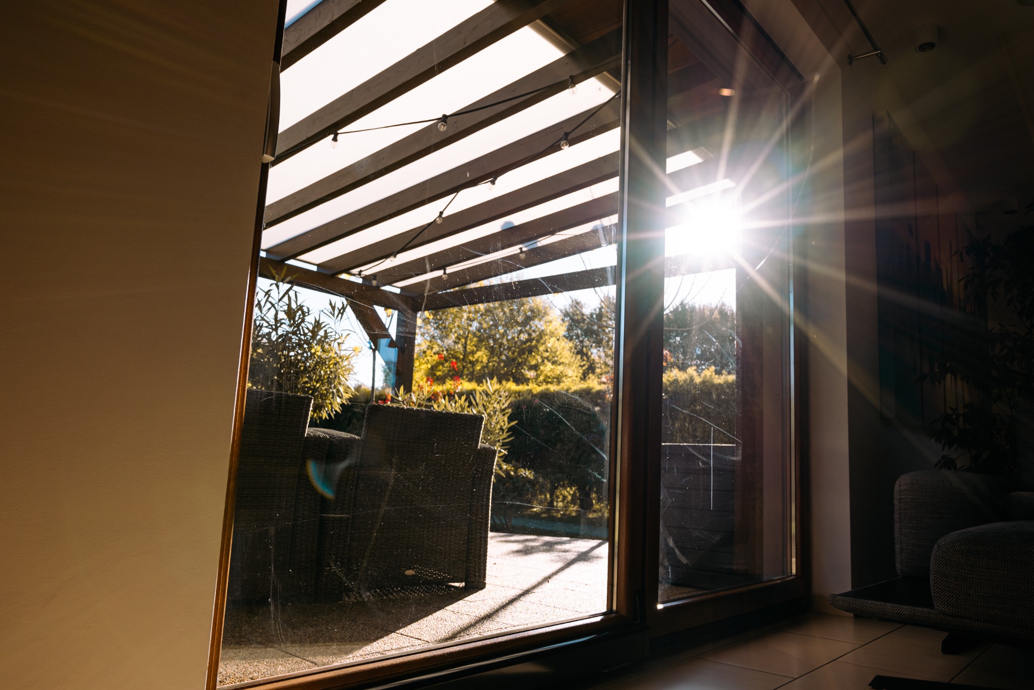 Przesuwne okno tarasowe z widokiem na zadaszony taras z meblami ogrodowymi w słoneczny dzień. Na szybie widoczne drobne zabrudzenia.