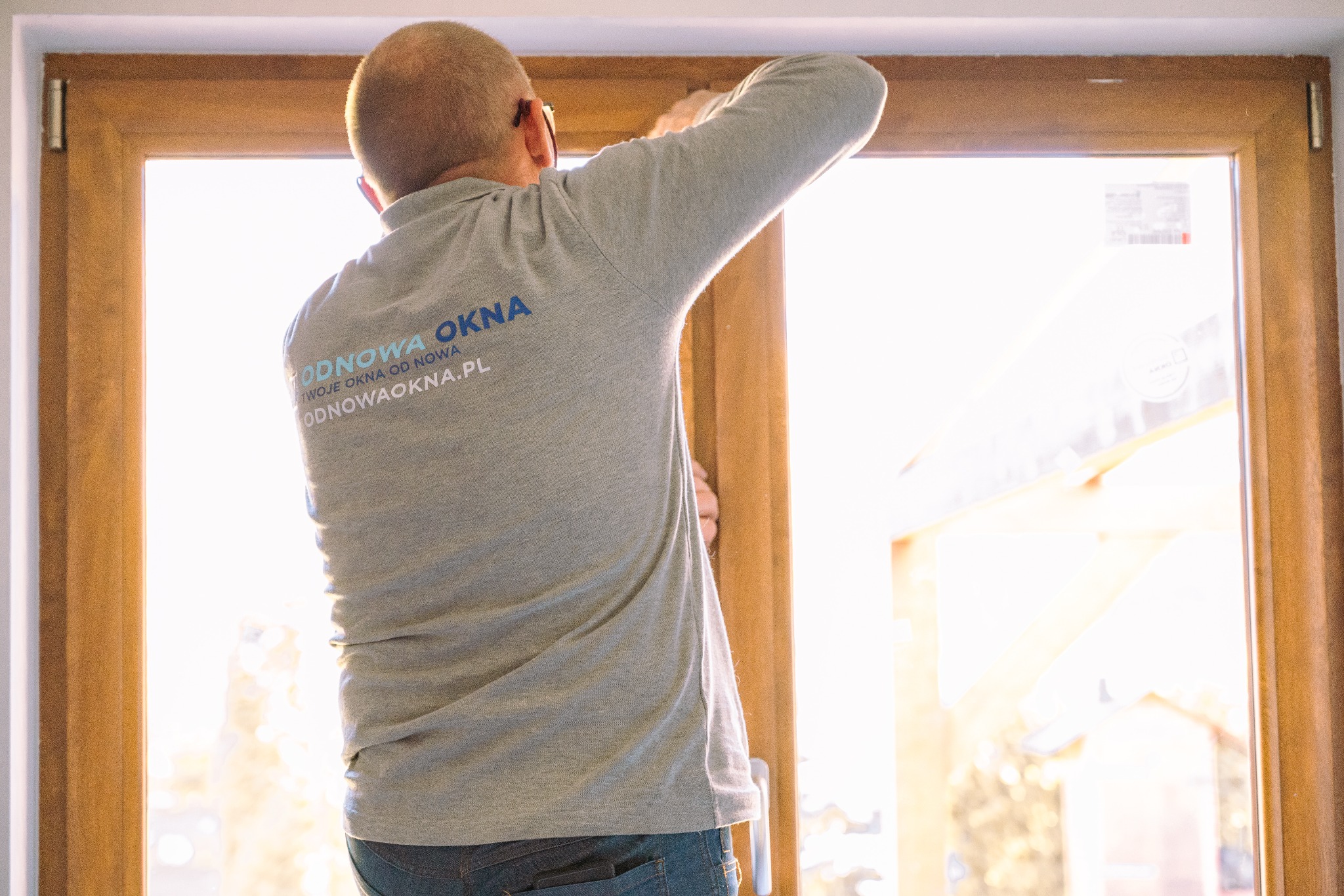 Mężczyzna w szarej koszulce z logo firmy montuje okno z drewnopodobną ramą, widok od tyłu, jasne światło słoneczne wpadające przez okno.