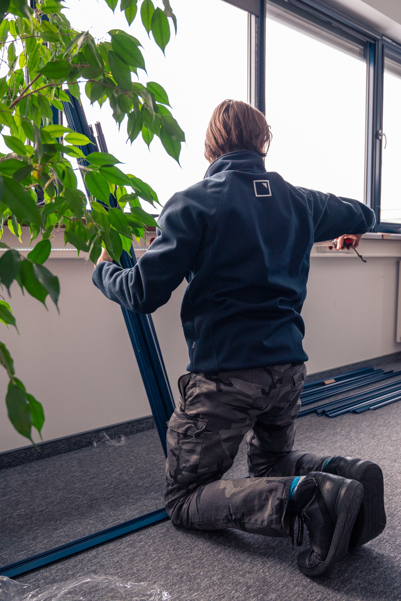 Instalator na kolanach montuje ramę okienną w pomieszczeniu biurowym, widoczne profile okienne, roślina doniczkowa i okno w tle.