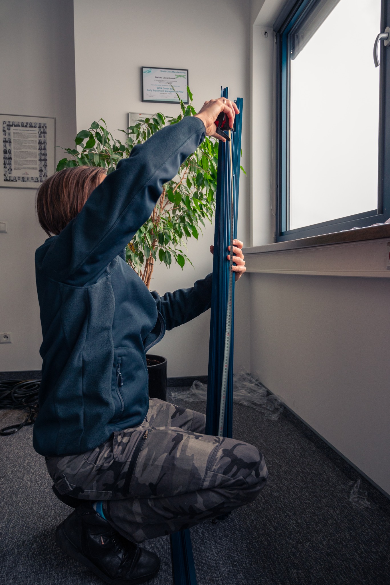Mężczyzna w pozycji kucznej mierzy niebieski profil okienny aluminiowy przy oknie za pomocą miarki, w tle widoczna roślina doniczkowa i dyplom na ścianie.