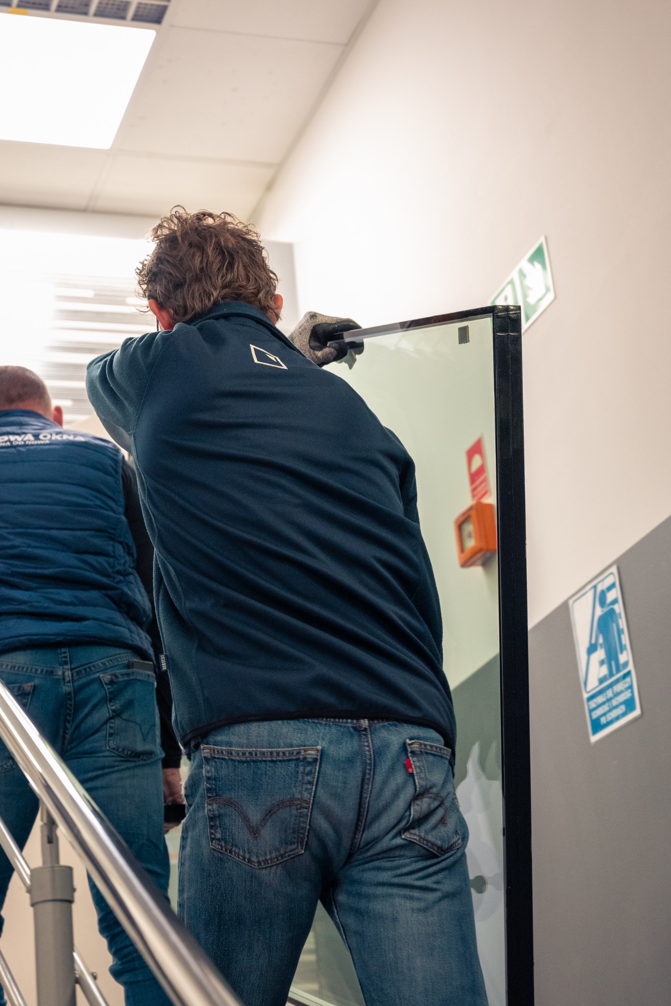 Pracownik w rękawicach ochronnych transportuje duże okno z czarną ramą po schodach wewnątrz budynku, widoczny fragment logo firmy na bluzie oraz tabliczki informacyjne na ścianie.