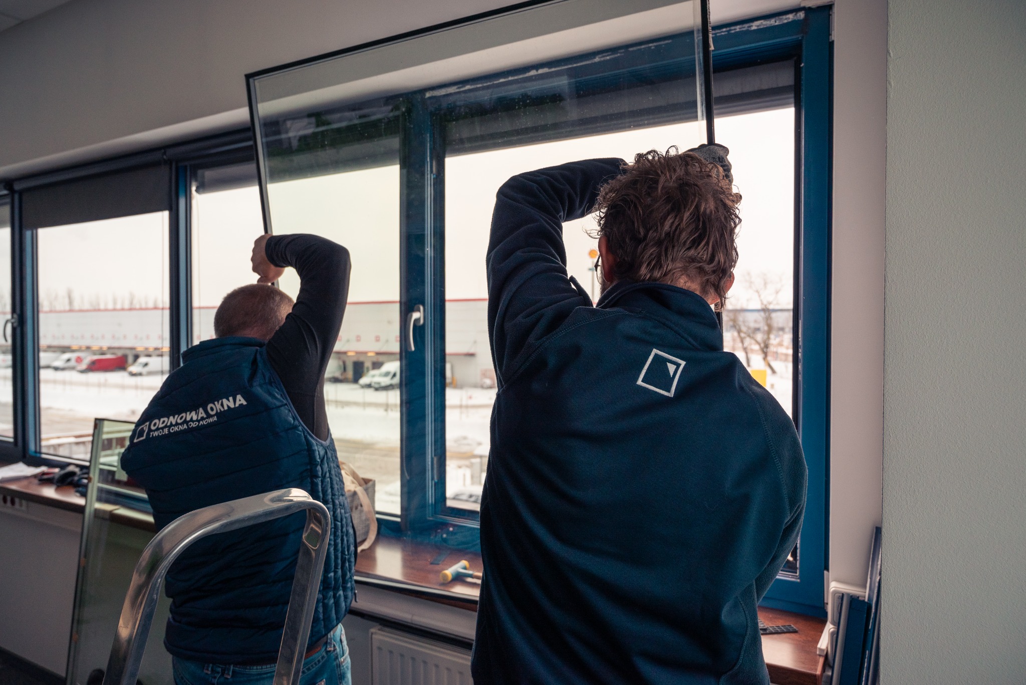 Dwóch monterów w uniformach instaluje szybę w ramie okiennej; widok zza pleców, w tle budynek magazynowy i drabina.