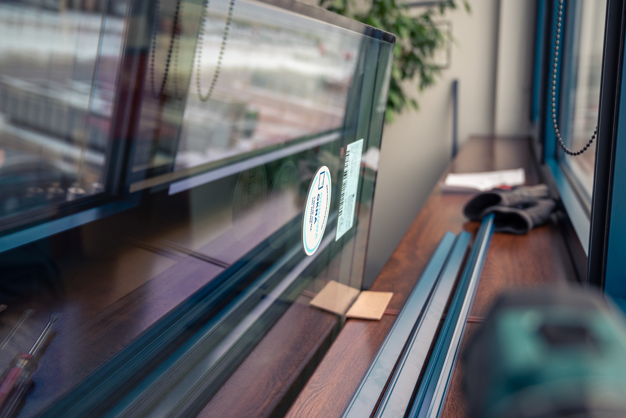 Przygotowane do montażu okno z naklejkami producenta, leżące na drewnianym parapecie obok narzędzi i rękawic roboczych.