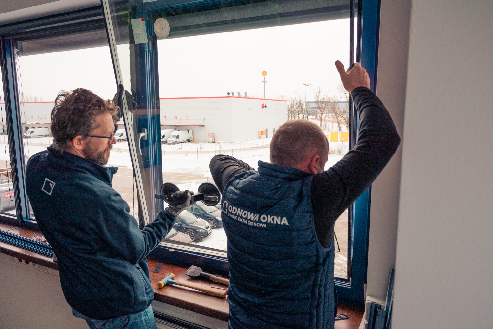 Dwóch monterów instaluje okno, używając przyssawek do szkła; widoczne narzędzia i logo firmy na kamizelce jednego z nich; za oknem zimowy krajobraz z budynkami magazynowymi.