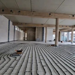 Wnętrze w stanie surowym z widoczną instalacją ogrzewania podłogowego, rury ułożone na macie systemowej z widocznymi napisami 'x-net' i 'ERMI', surowy betonowy strop z drewnianymi belkami...