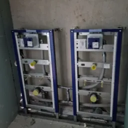 Dwa moduły podtynkowych stelaży WC z niebieską ramą zamontowane na surowej ścianie, widoczne rury i elementy instalacji.