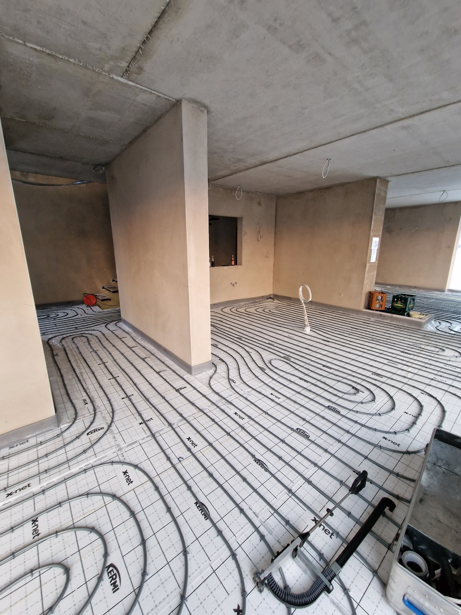Rozłożona instalacja ogrzewania podłogowego z rurami Kermi X-net na białej macie w surowym wnętrzu z betonowym stropem, widoczne narzędzia i skrzynka instalacyjna.