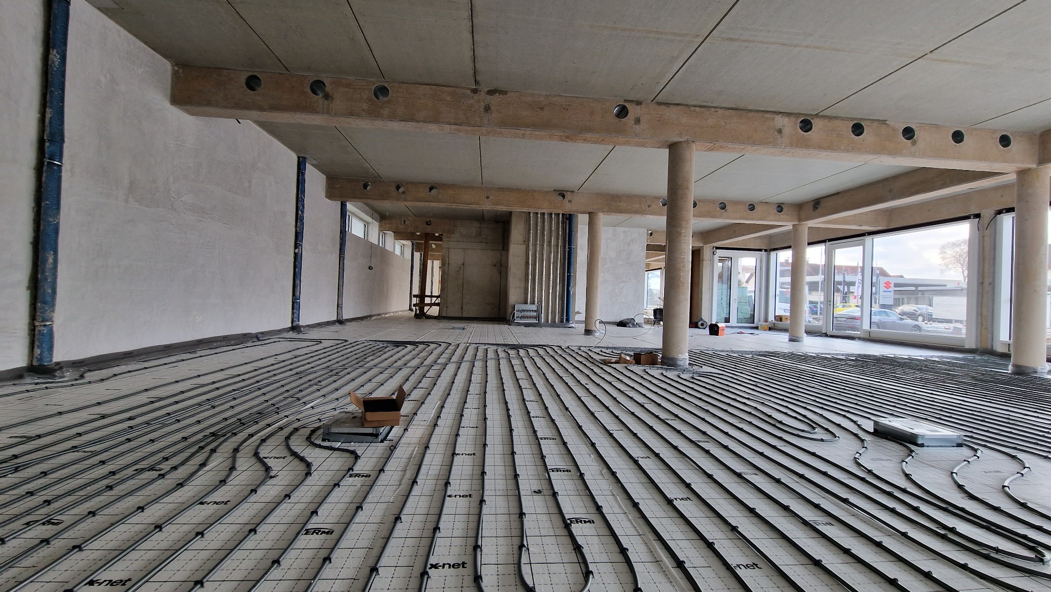 Wnętrze w stanie surowym z widoczną instalacją ogrzewania podłogowego, rury ułożone na macie systemowej z widocznymi napisami 'x-net' i 'ERMI', surowy betonowy strop z drewnianymi belkami...