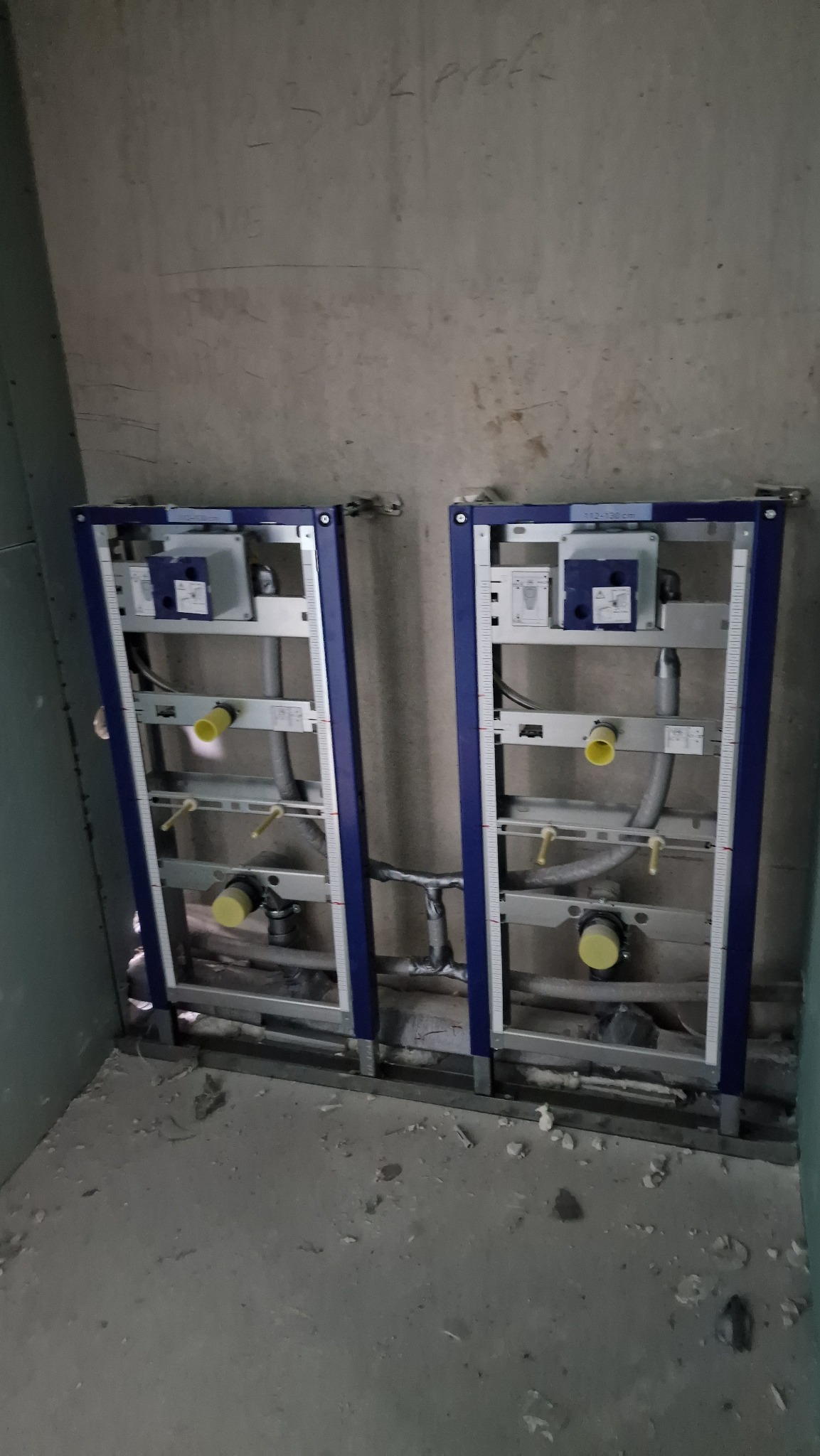 Dwa moduły podtynkowych stelaży WC z niebieską ramą zamontowane na surowej ścianie, widoczne rury i elementy instalacji.