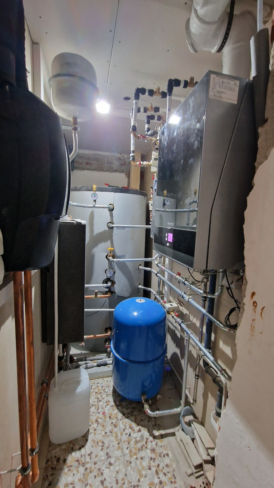 Kotłownia w trakcie montażu: widoczny nowy piec gazowy, zbiornik buforowy, naczynie przeponowe w kolorze niebieskim oraz liczne rury miedziane i plastikowe. Ściany częściowo wykończone płytami...