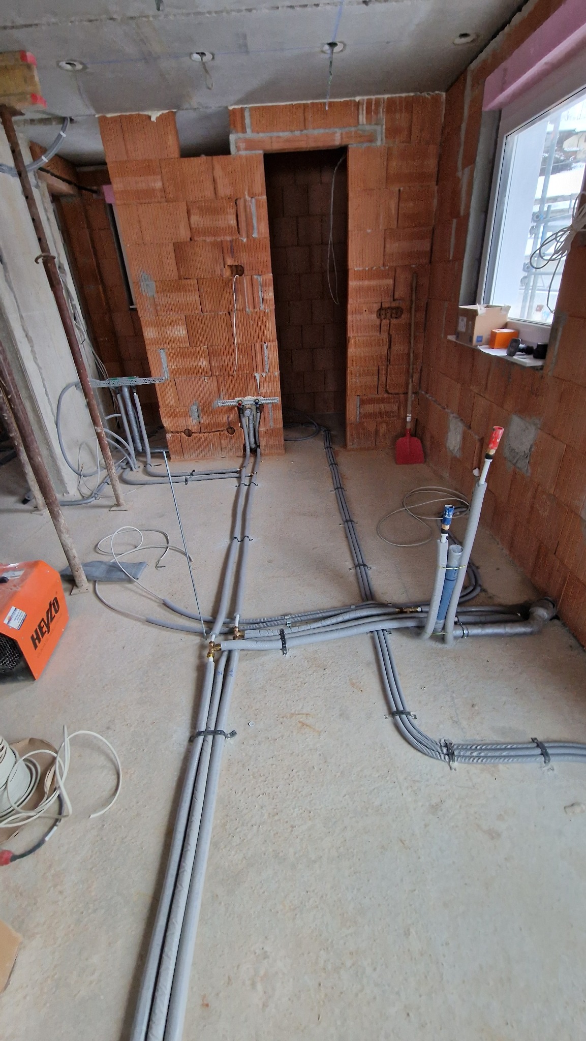 Szare rury instalacyjne prowadzone po podłodze w surowym wnętrzu z ceglanymi ścianami i oknem w tle, widoczny grzejnik elektryczny.