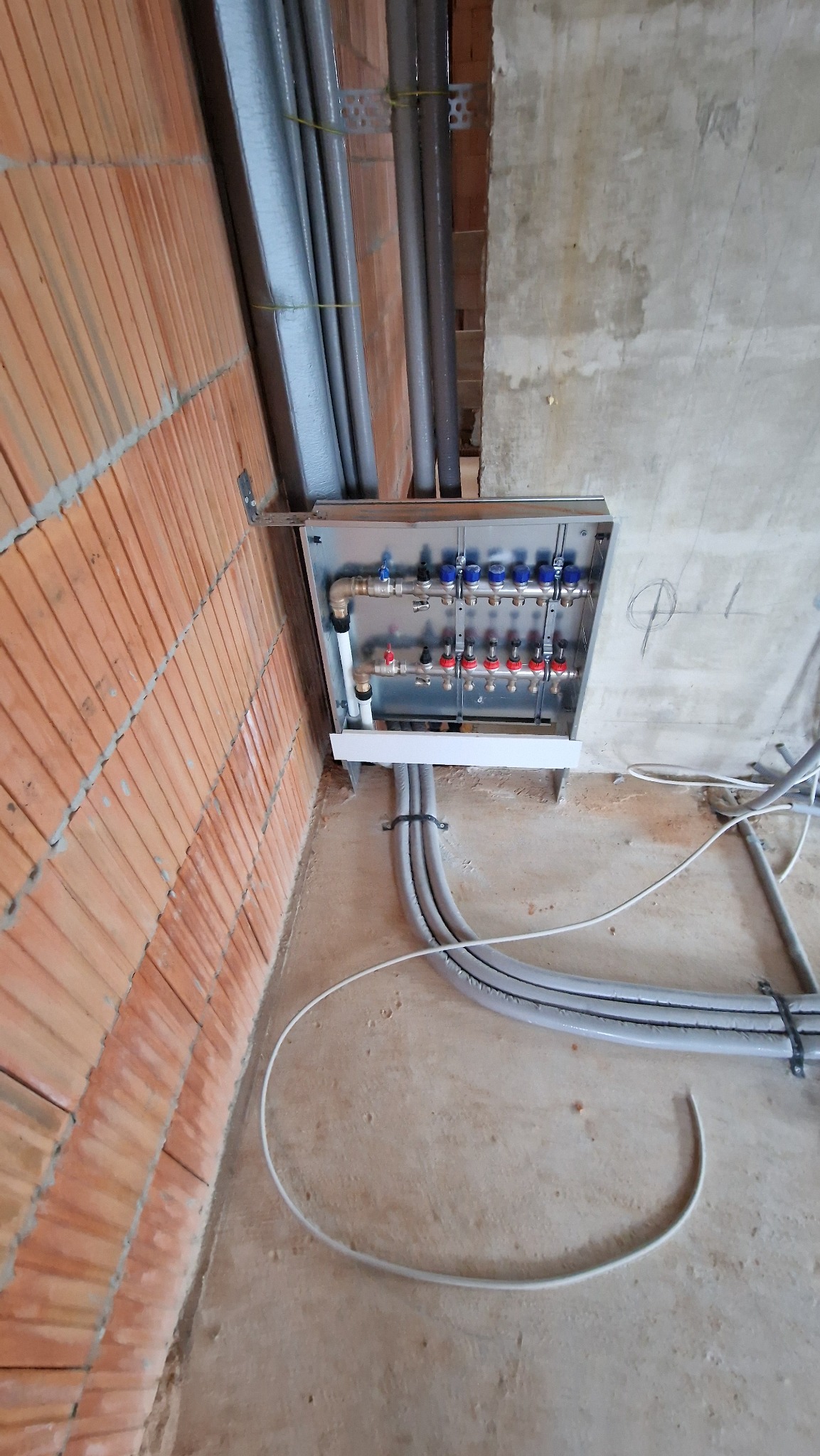 Rozdzielacz ogrzewania podłogowego w metalowej szafce, z widocznymi rurami doprowadzającymi i powrotnymi, zamontowany na surowej ścianie z cegły i betonu.