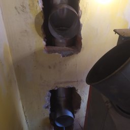 RP-Kom Frezowanie Rozwiercanie Kominów - Dwa otwory w żółtej ścianie z wystającymi rurami kominowymi ze stali nierdzewnej, widoczne fragmenty cegieł i resztki tynku wokół otworów, z boku fragment pieca.