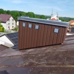 Brązowy komin wentylacyjny z trzema metalowymi kratkami na dachu z blachy, w tle budynki mieszkalne i kościół w słoneczny dzień.