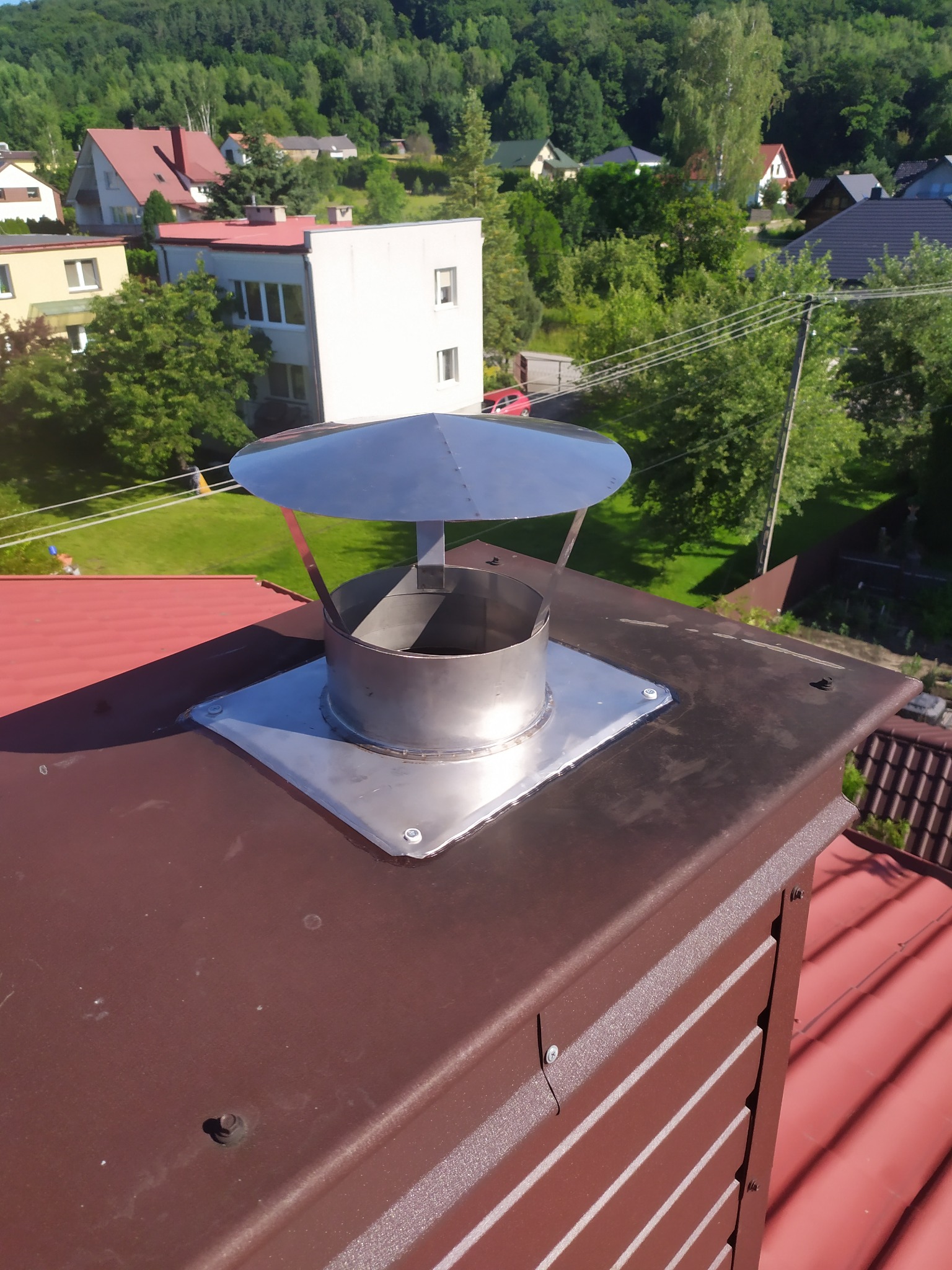 Błyszcząca, metalowa nasada kominowa z daszkiem zamontowana na brązowym kominie z blachy na tle zieleni drzew i budynków.