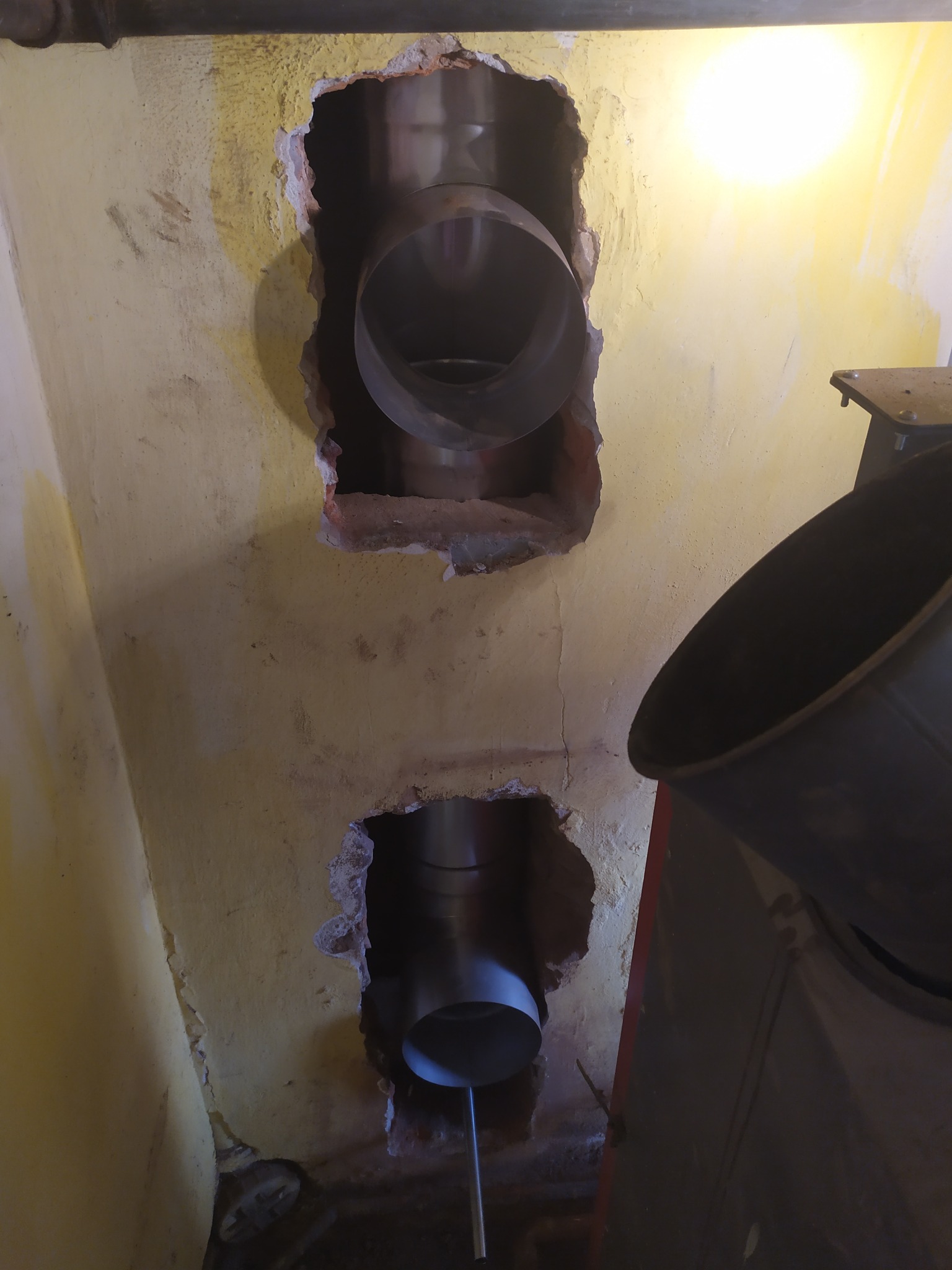 Dwa otwory w żółtej ścianie z wystającymi rurami kominowymi ze stali nierdzewnej, widoczne fragmenty cegieł i resztki tynku wokół otworów, z boku fragment pieca.