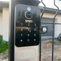 SmartMP Michał Polak - Nowoczesny wideodomofon z klawiaturą numeryczną i kamerą, zamontowany na czarnym słupku ogrodzenia panelowego, z widocznym logo producenta S-Tech.