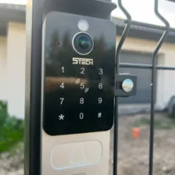 Nowoczesny wideodomofon z klawiaturą numeryczną i kamerą, zamontowany na czarnym słupku ogrodzenia panelowego, z widocznym logo producenta S-Tech.