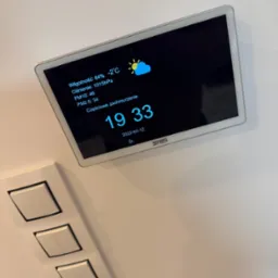 Panel sterowania inteligentnego domu na ścianie, wyświetlający dane o wilgotności, temperaturze, ciśnieniu, poziomie PM10 i PM2.5 oraz godzinę i datę, umieszczony obok włączników światła.