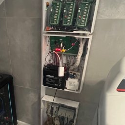 SmartMP Michał Polak - Rozbudowana centrala alarmowa w białej obudowie, z widocznymi płytkami drukowanymi, przewodami i akumulatorem Alarmtec, podłączona do laptopa wyświetlającego dane konfiguracyjne.